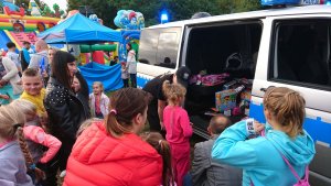 Zdjęcie ukazuje dzieci z opiekunami czekające na upominki, które to wręcza policjant ze swojego busa policyjnego. W tle widoczny jest dmuchany zamek dla dzieci.