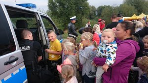 Zdjęcie ukazuje dzieci z opiekunami czekające na upominki, które to wręcza policjant ze swojego busa policyjnego. W tle widać inne atrakcje oraz policjanta z motocyklem policyjnym.
