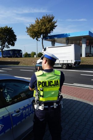 Na zdjęciu widnieje policjant stojący obok radiowozu przy drodze, trzymając w ręce fotoradar, dokonuje rutynowej kontroli prędkości na drodze.