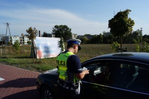 Zdjęcie przedstawia policjanta, który stojąc przy zatrzymanym przez niego samochodzie, dokonuje kontroli dokumentów pojazdu oraz kierowcy.