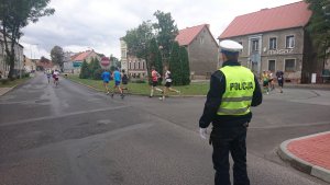 Zdjęcie przedstawia biegaczy uczestniczących w biegu a także policjanta, który zabezpiecza trase biegu w mieście.